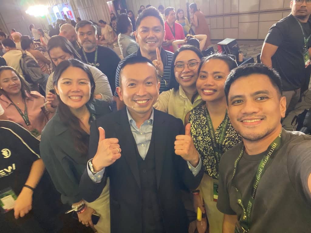 Attendees wearing event lanyards smile for a group selfie, with thumbs up in the foreground.