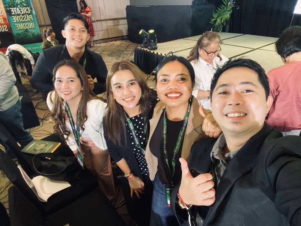 Five attendees wearing event lanyards pose for a group selfie near the stage.